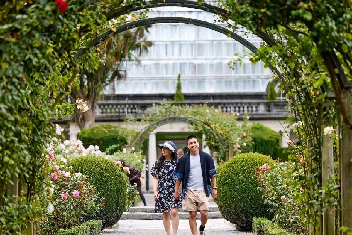 A candid photo of a romantic couple walking hand-in-hand through a lush rose garden archway at a scenic wedding venue.