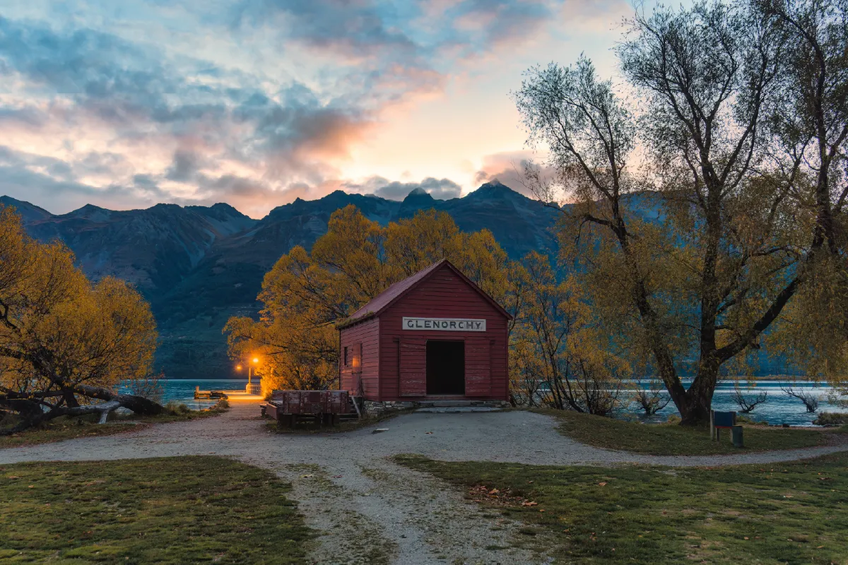 The historic Glenorchy red shed at twilight, featuring soft sunset colors and glowing wharf lights by the lake and mountains.