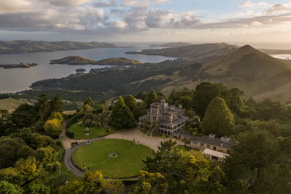 An aerial drone shot of Larnach Castle in Dunedin, showing the grand estate and garden overlooking the harbor at sunset.