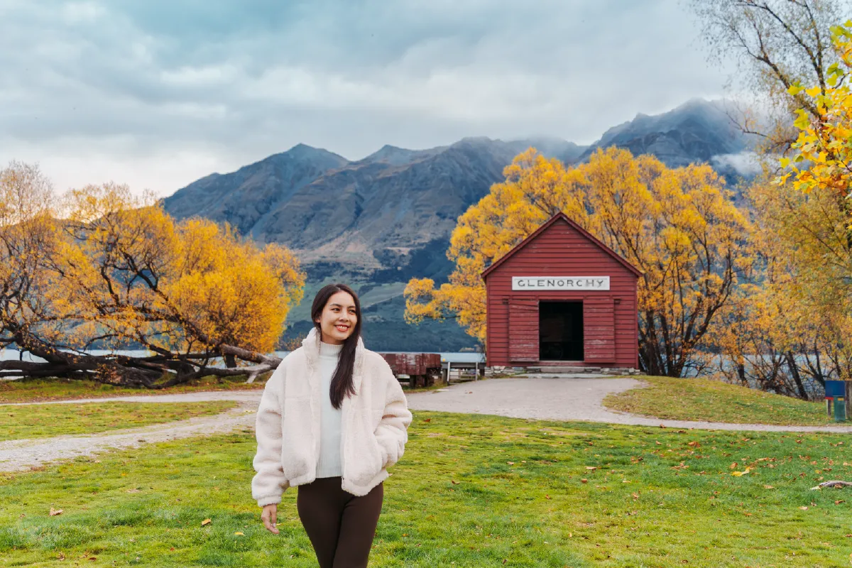 A woman smiling in front of the iconic red Glenorchy Steamship Depot with golden autumn trees and mountains in the background.