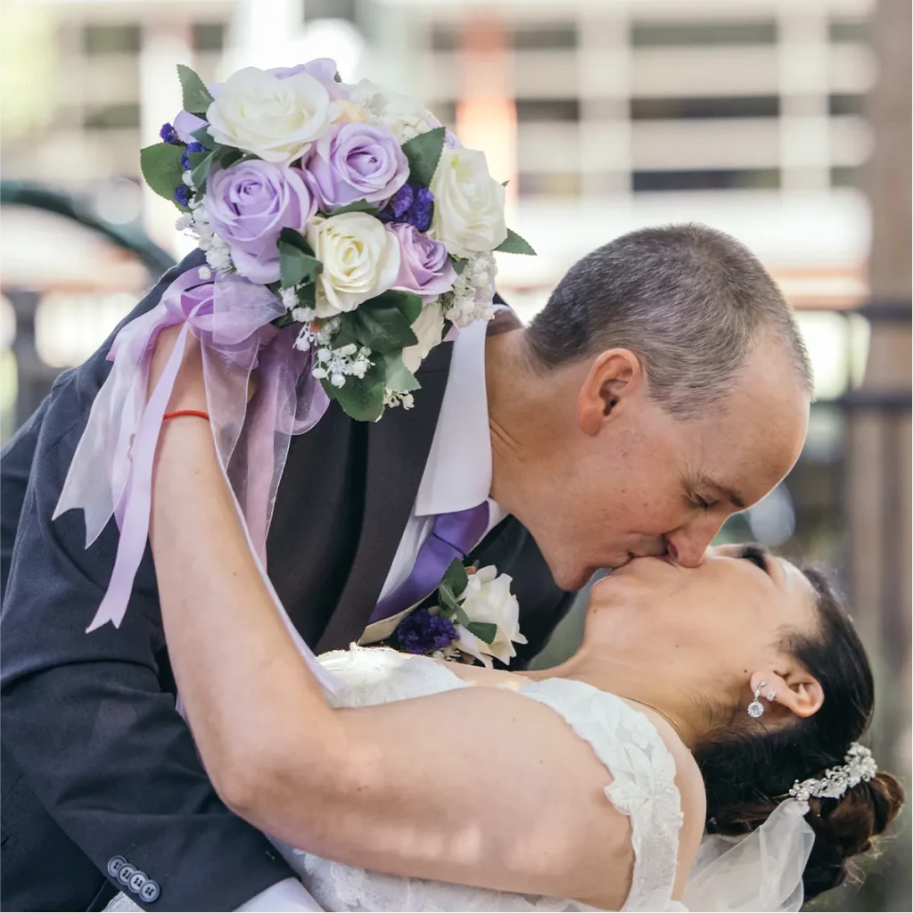 A groom dipping and kissing his bride while she holds a purple and white rose bouquet.