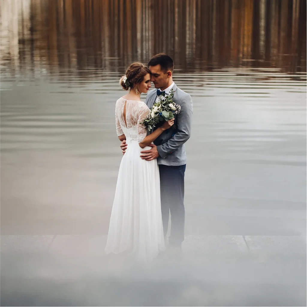 A bride and groom standing on a misty dock with calm water and reflections behind them.