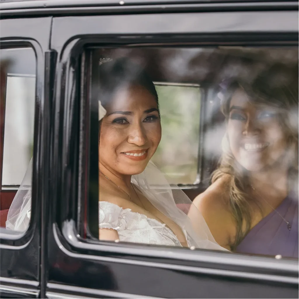 A bride smiling from the back of a vintage black wedding car with her bridesmaid.
