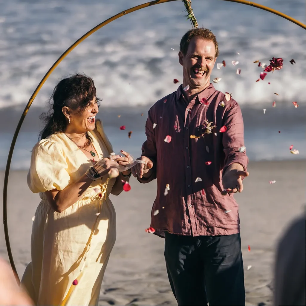 A joyful couple laughing as rose petals are thrown during a beach wedding ceremony.