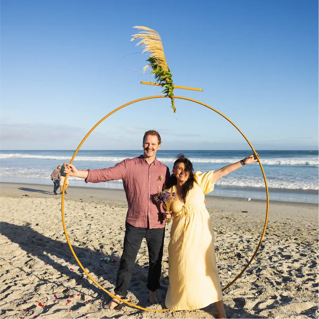 Newlyweds posing inside a circular gold wedding arch on a sunny beach in New Zealand.
