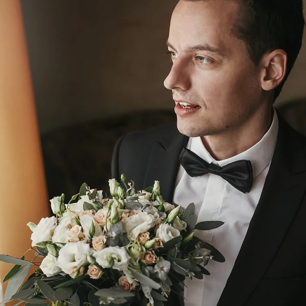Close-up of a groom in a black tuxedo and bowtie holding a lush bridal bouquet.